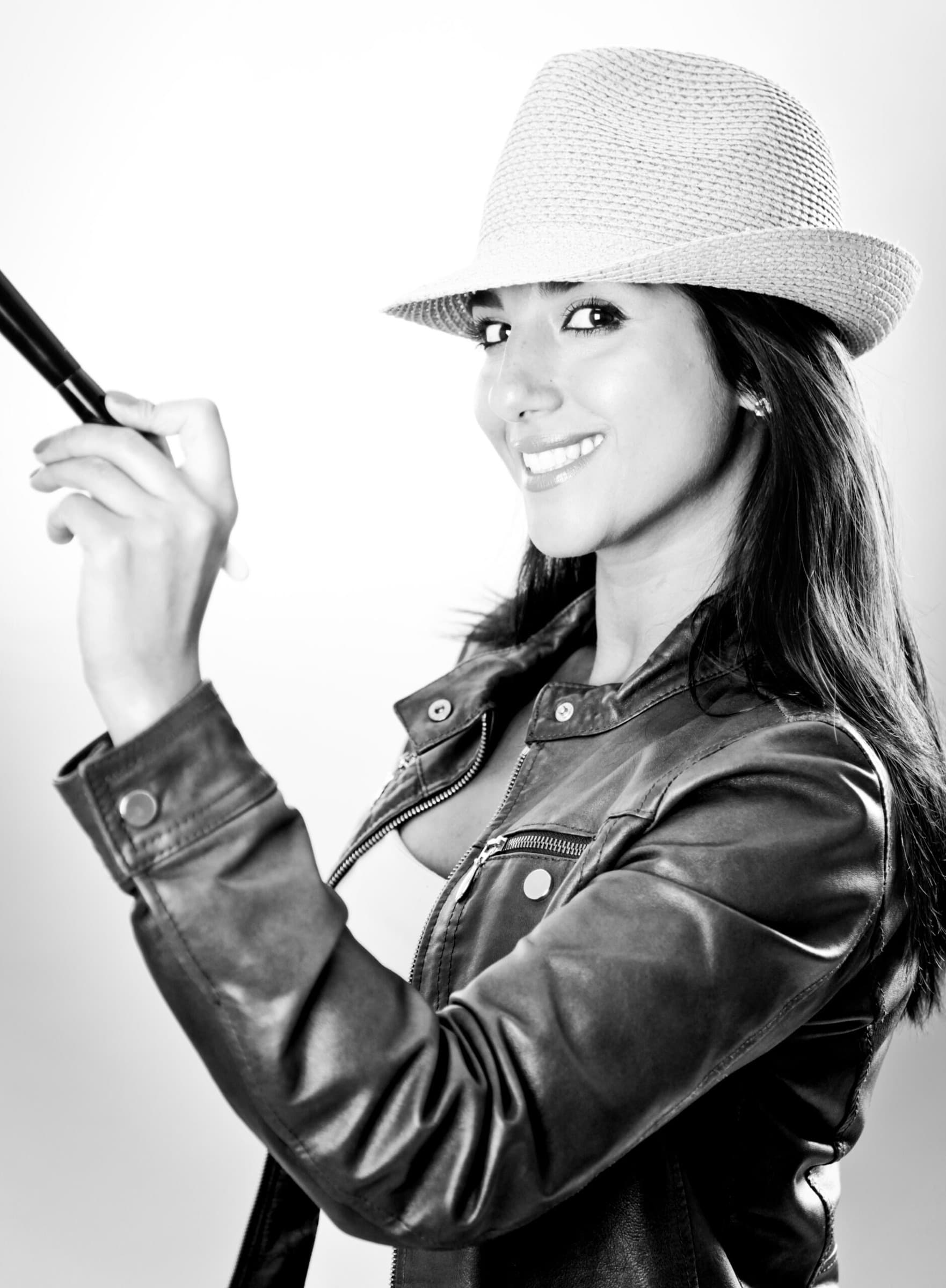 Schwarzweiss-Porträt einer Frau mit Hut und Lederjacke, die einen Make-up-Pinsel hält